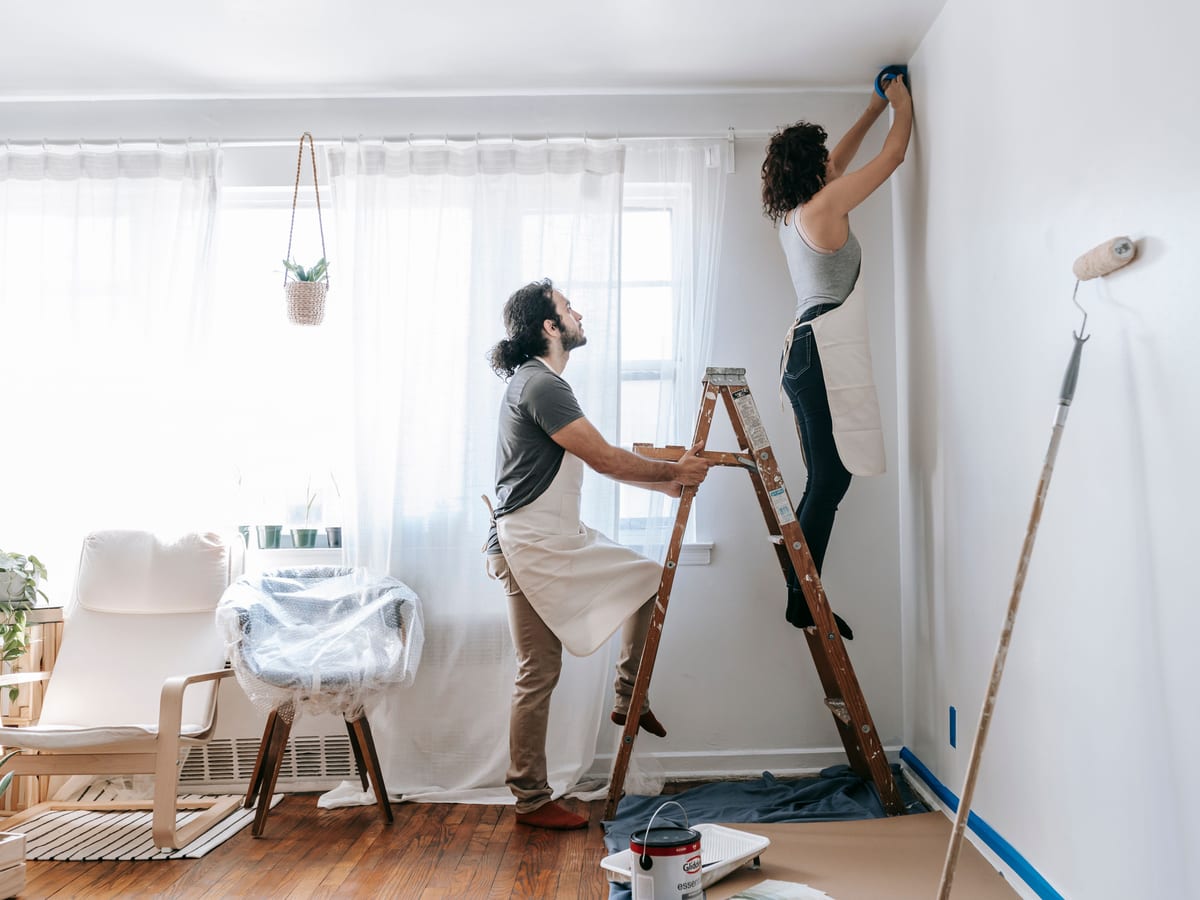 Couple renovating their home, with man supporting ladder and woman working on walls indoors.